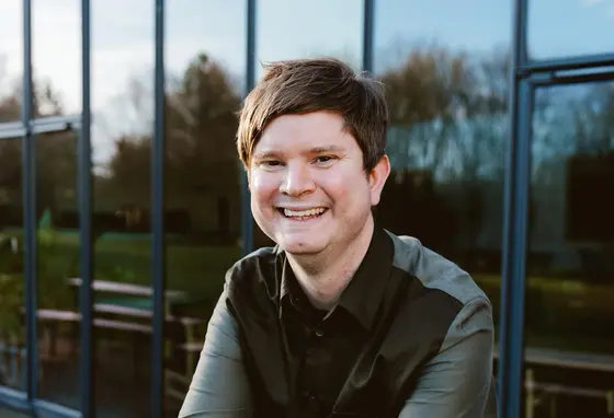 a photo of David Sheppeard sitting outside against a glass wall. David has white skin, short brown hair and wears a dark shirt