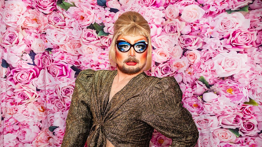 A drag performer wearing styled blond hair, exaggerated make-up and a metallic gold top against a colourful floral backdrop