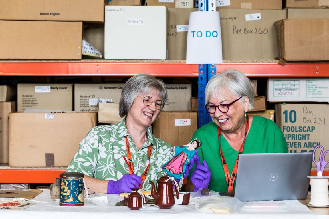 Barbara and Fiona, two smiling women both with white hair and glasses, sit at a desk in the open store and examine pottery and homewares from the collection.