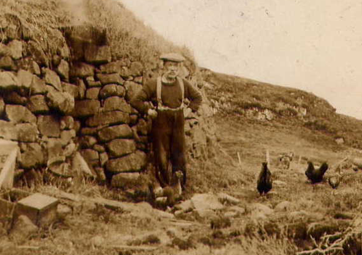 Historic photograph showing someone standing outside a stone house. Two chickens are around the persons feet.