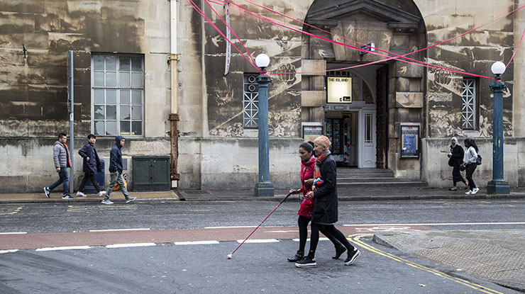 Two women cross a street, one with a stick.