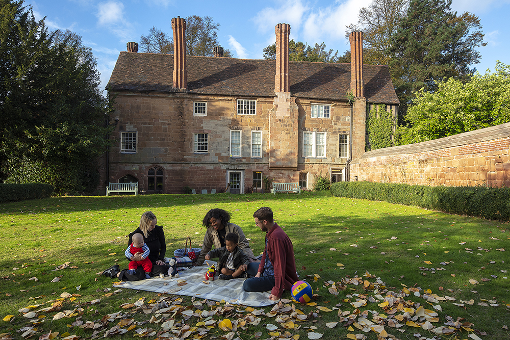 Five people sit on a blanket outside a stately house