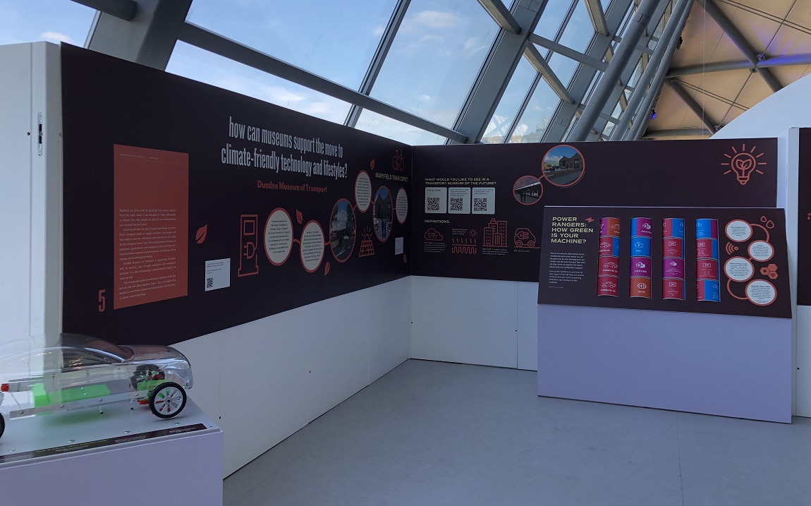An exhibition space with panels of information, imagery and a model electric car in the corner