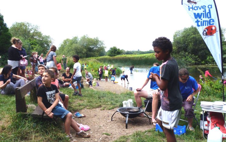 A group of children and adults are sitting and standing by a river doing nature-based activities.