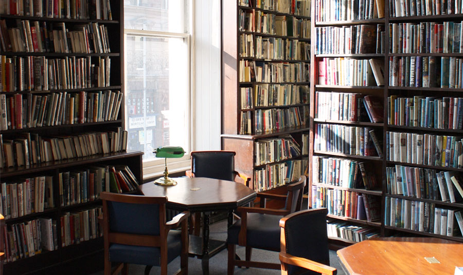 Sun shining through a library window in between book shelves. Tables and chairs are in the foreground.