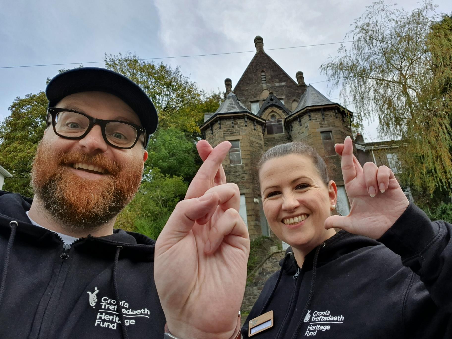 A man and a woman are stood in front of a heritage site doing the crossed fingers selfie