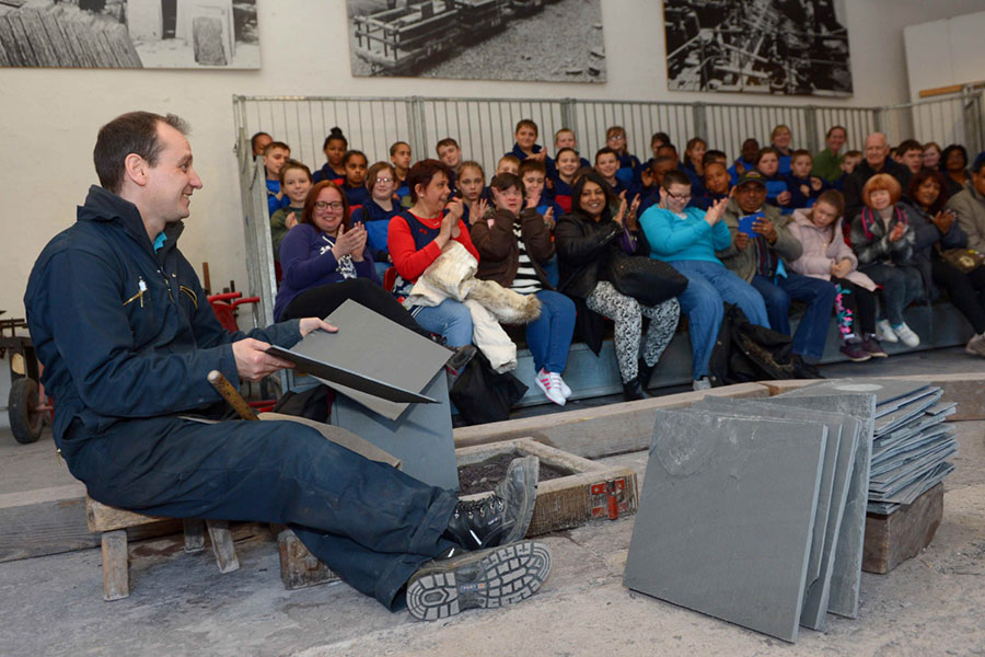A man givign a demonstration of slate cutting in front of a large seated crowd