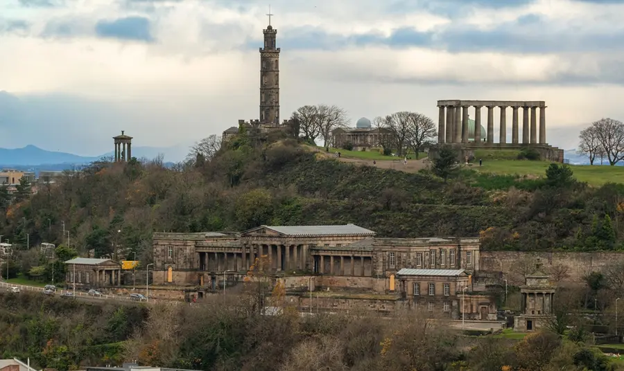 A skyline view of Calton Hill in Edinburgh. In the foreground, the Royal High School stands on the steep banks of the hill. At the summit, the Nelson Monument and the National Monument for Scotland stand tall against the sky.