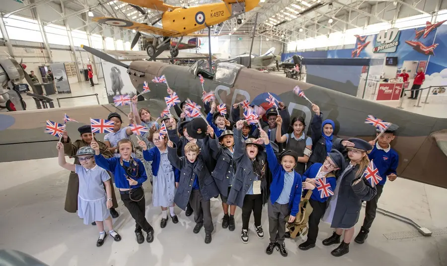 A group of schoolchildren mark the 80th anniversary of VE Day during a visit to RAF Museum Midlands. A group of schoolchildren shout and wave union flags in front of aircraft from the museum's collection.