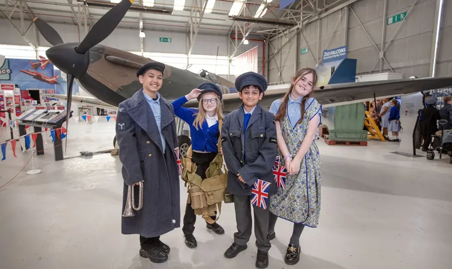 Young visitors mark the 80th anniversary of VE Day during a visit to RAF Museum Midlands. Four children, dressed in WW2 attire and holding union flags, stand in front of a Spitfire at the RAF Museum Midlands.