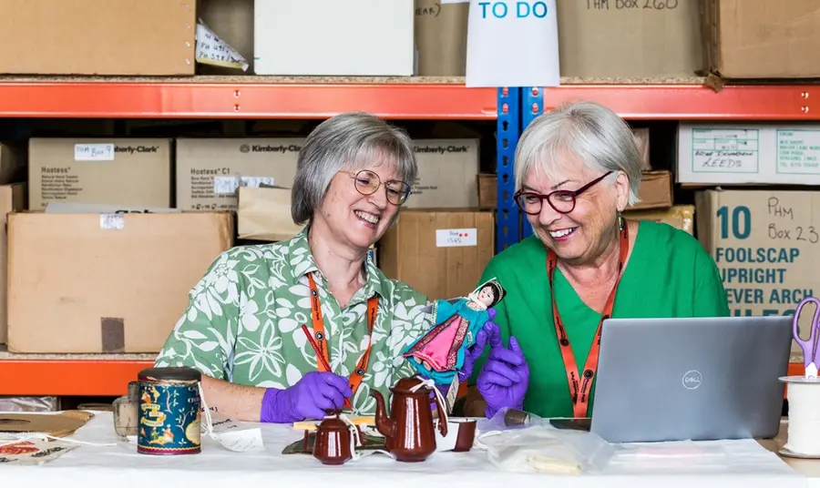 Barbara and Fiona sit at a desk in a collections store, smiling as they assess pottery and porcelain items from the collection.