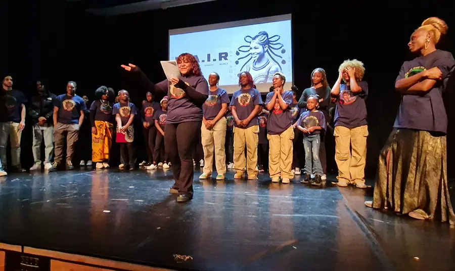 A large group on stage with a person reading from a sheet of paper in the foreground