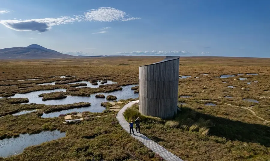 A remote, contemporary bird-watching tower in the middle of vast wetlands. Across a wooden causeway, two birdwatchers depart the tower.