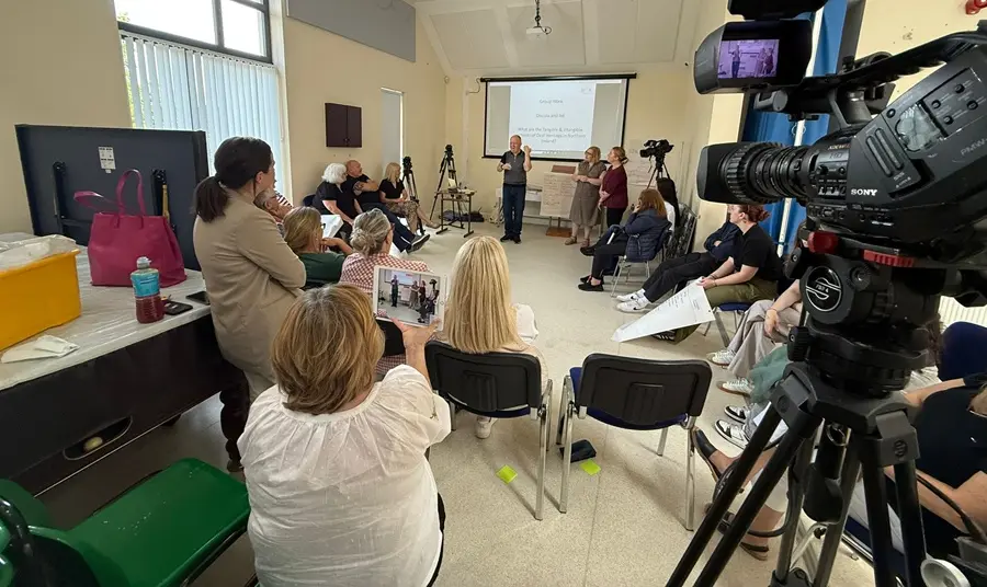 People sit on chairs in a circle watching someone give a presentation. In the foreground a video camera films them.