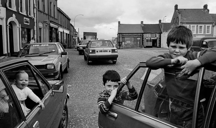 Two young boys stand around an open car door to the right. On the left, a baby is held up through the open window of another parked car.