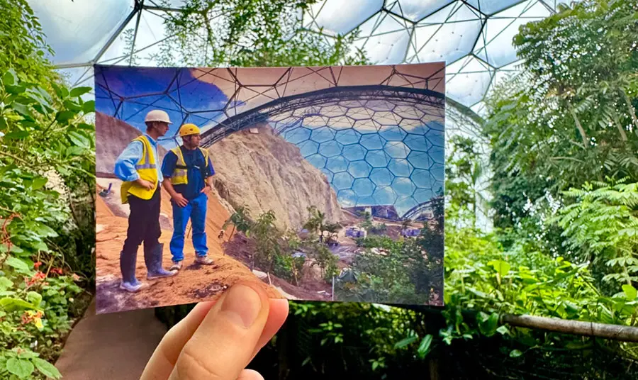 A hand holding a photograph of two people standing inside a geodesic dome, with that same geodesic dome in the background