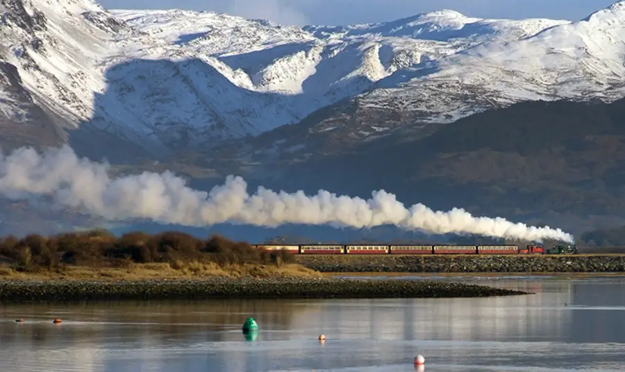 A steam train travelling next to water with snowy mountains behind