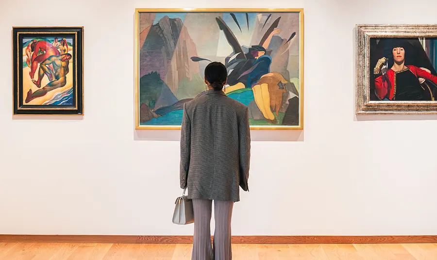 A young person looking at one of three paintings on a gallery wall. The person has their back to us.