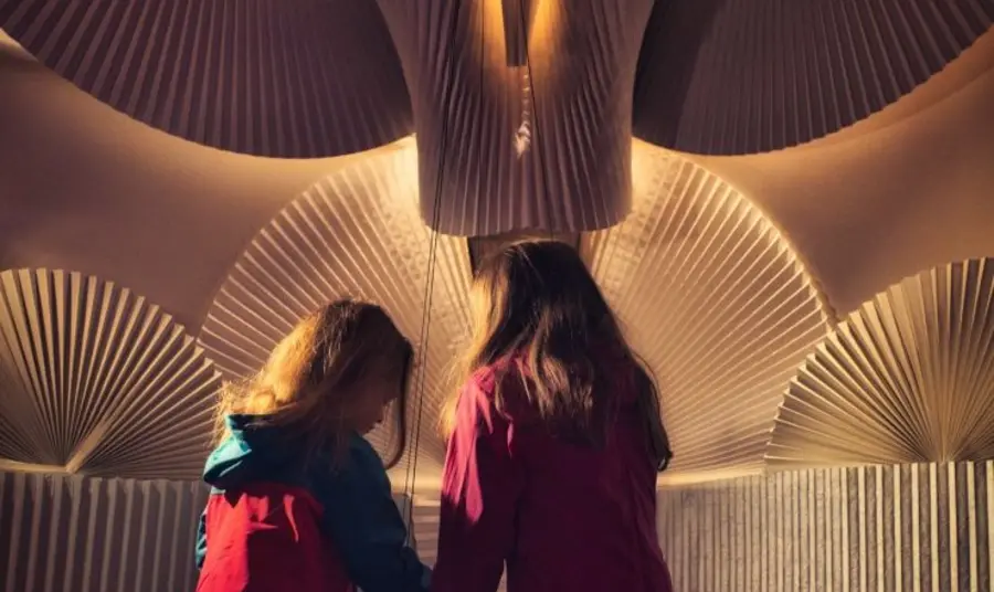 Two children play with the sand pendulum exhibit. The backdrop is made from reclaimed window blinds arranged into patterns