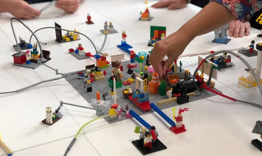 A structure made of Lego in the middle of a table with people sat around it, contributing to building it