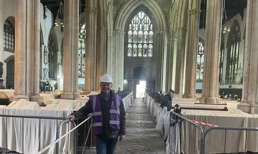 A smiling man, wearing a hard hat and a purple high visibility jacket, stands in the central nave of the church, where the pews are covered in dust sheets and protective barriers.