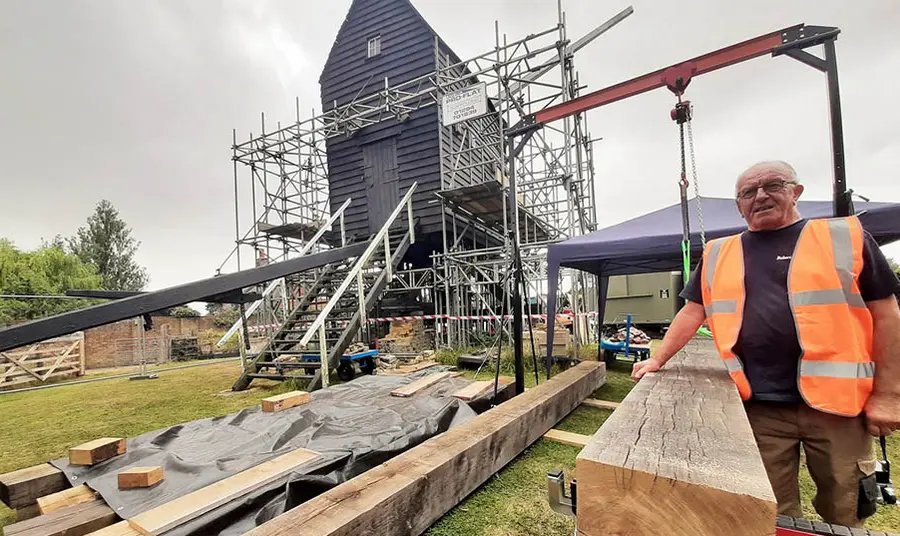 A construction worker standing next to a windmill covered in scaffolding with large wooden beams on the ground.