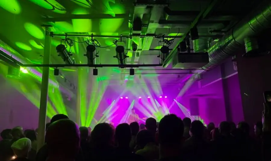 The view from the back of the room shows a crowd of people and a concert stage lit up with vibrant colours