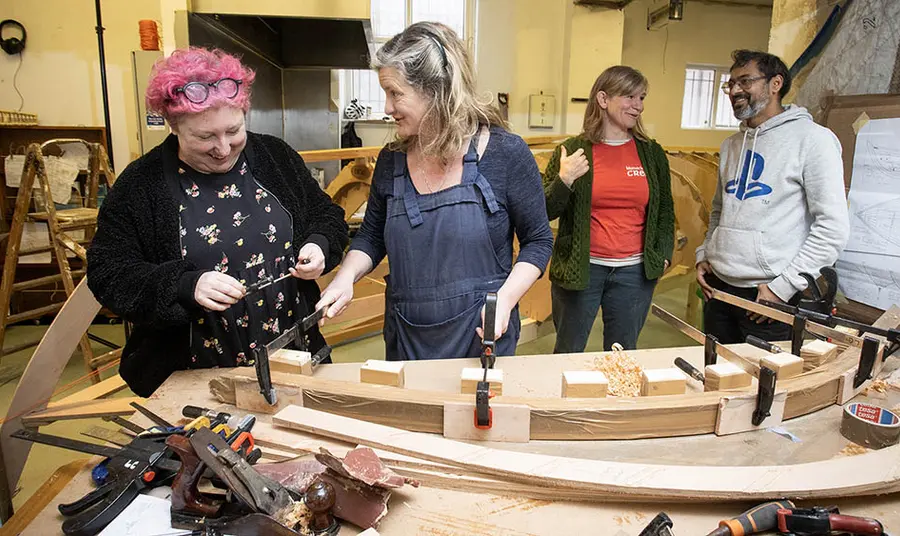 Four people stand in a workshop with boatbuilding equipment on a table in front of them