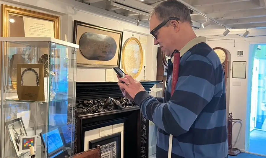 A person holding a white cane and using a smartphone, in front of them is a glass museum case containing old photographs and a brightly coloured NaviLens code