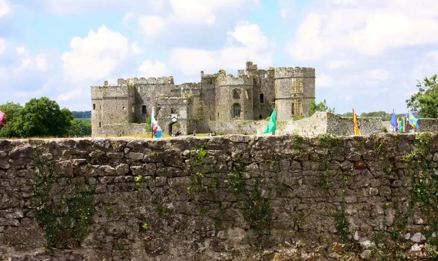 A centuries old derelict castle behind a dry stone wall, with visitors and flags outside on the grounds.
