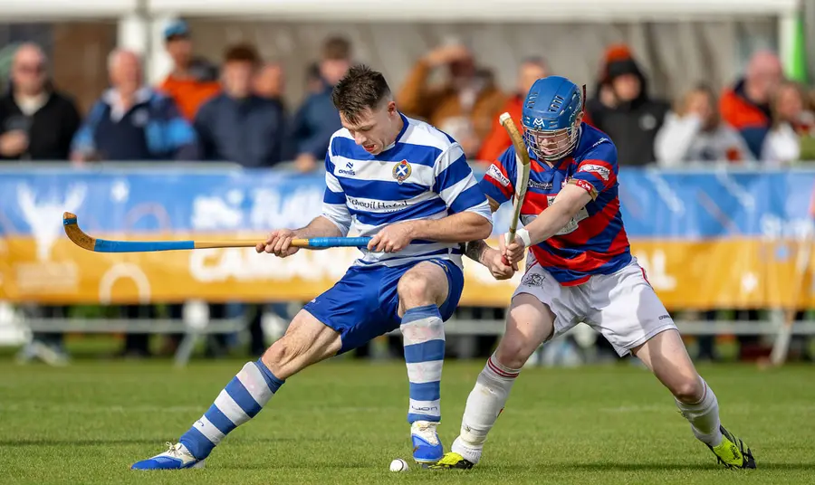 two shinty players one in blue and white the other in red and blue tustle over a ball