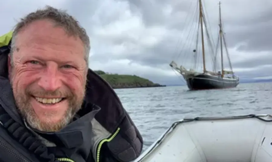 Clive, smiling and wearing a life jacket while at sea on a dinghy, shares a view of a tall ship at sail behind him.