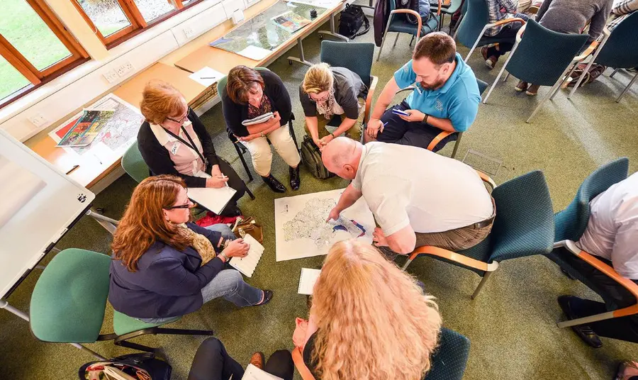 A group of people sit in a circle of chairs, pointing out and discussing details of a map of Cheshire on the floor between them.