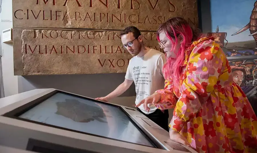 Two people using a digital display screen in a museum, with a large carved stone behind them