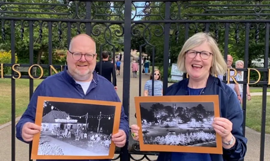 David and Carole, smiling, stand outside the gates of Jephson Gardens, the historic venue for the light show, holding black and white photographs of the original exhibitions.