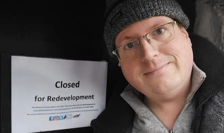 David, wearing glasses and a woollen hat, stands by a door bearing the sign 'closed for restoration'.