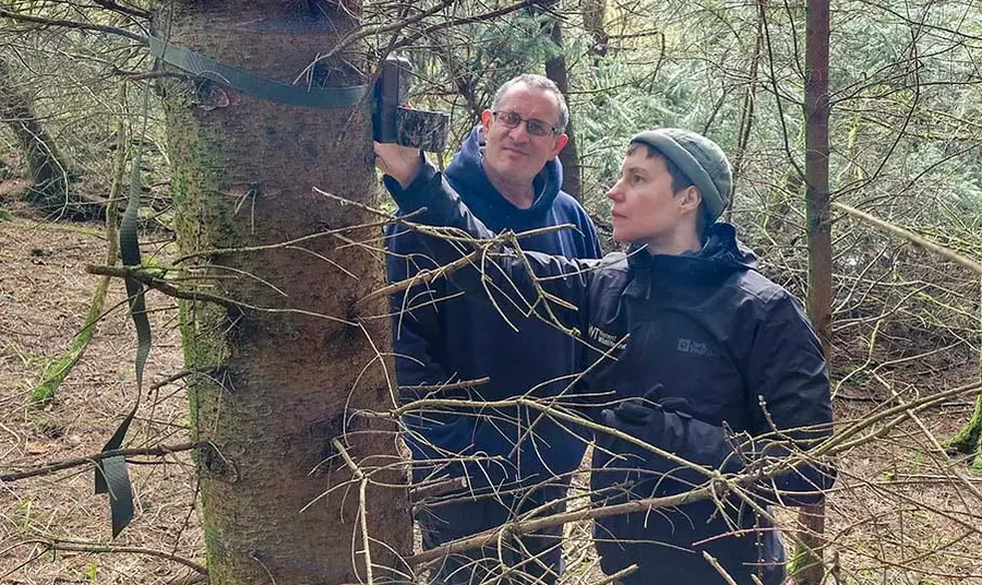 Two volunteers wearing outdoor gear stand in a pine forest an look at a den box which has been strapped to a tree