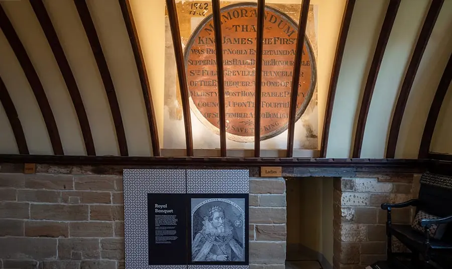 A photo of a wall and part of a ceiling inside a building, showing a large plaque commemorating a visit to the building by King James I in 1617