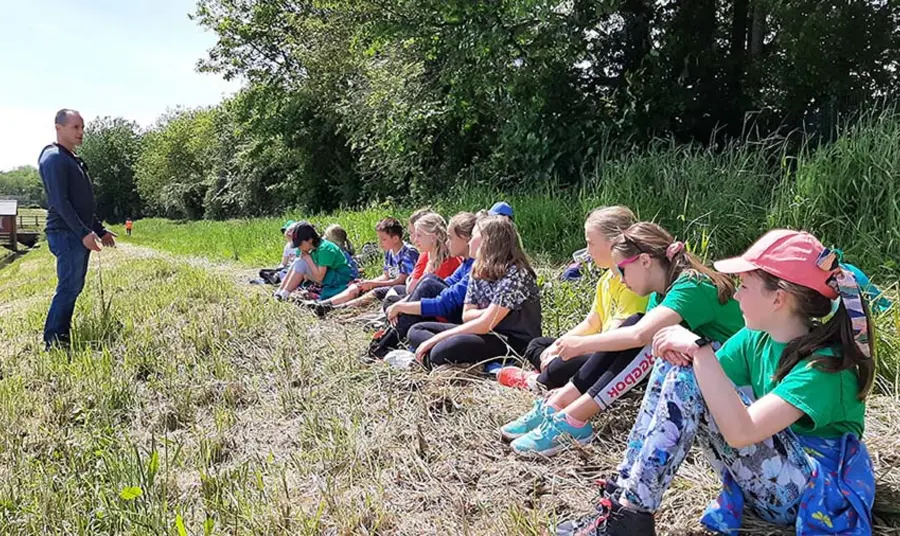 Plant yn eistedd ar laswellt ar ymyl coetir yn gwrando ar oedolyn