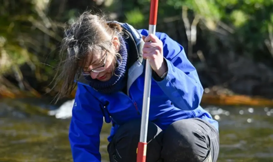 Elizabeth, wearing a blue rain coat and wellington boots, crouches in a shallow river and uses a net to sift through the current.