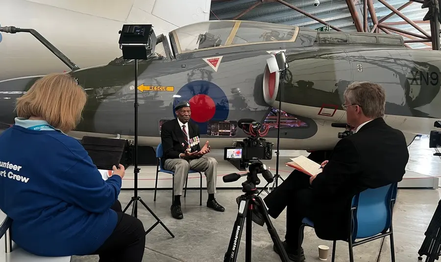 A veteran, sitting in front of a fighter jet in the RAF Museum, shares his testimony with a film crew and interviewer.