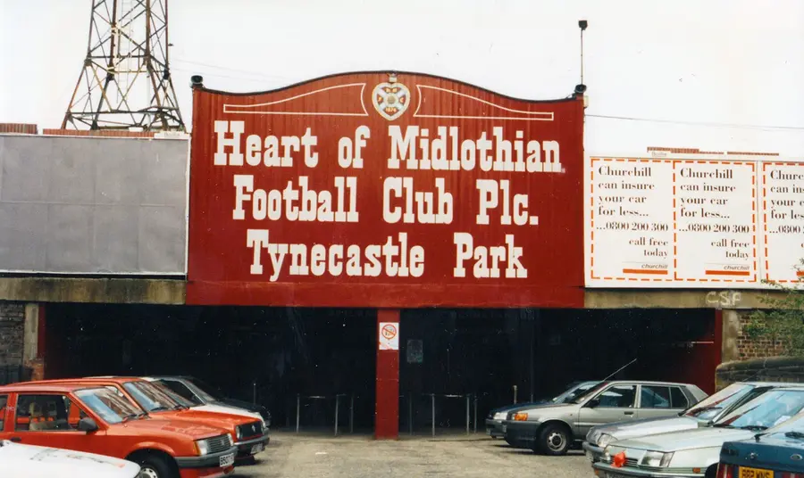 The gates to Tynecastle Park, the Hearts' stadium, in the 1970s or 80s. Cars of the era are parked in the foreground, and a large sign above the gates declares the name of the club and the stadium.