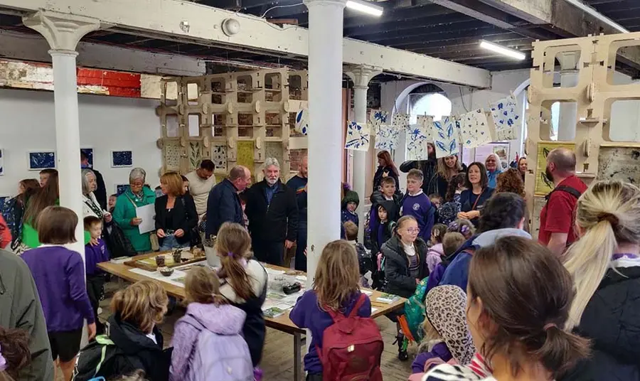 A group of people including children wearing school uniforms inside a historic industrial building with large white metal beams and columns