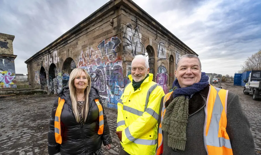 Three people wearing high vis jackets stand in front of a derelict historic building in a post-industrial landscape, formerly a shipyard.
