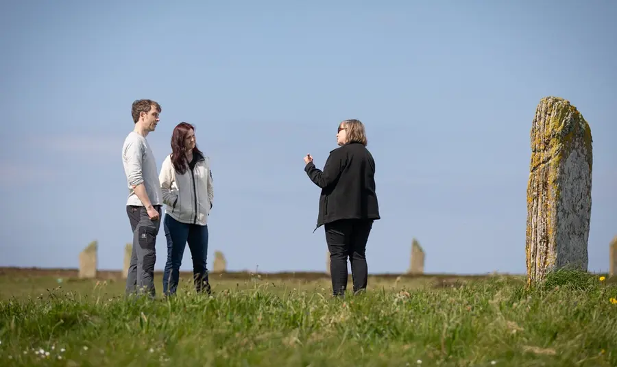 Two visitors listen to a tour guide explain the history of a ring of standing stones.