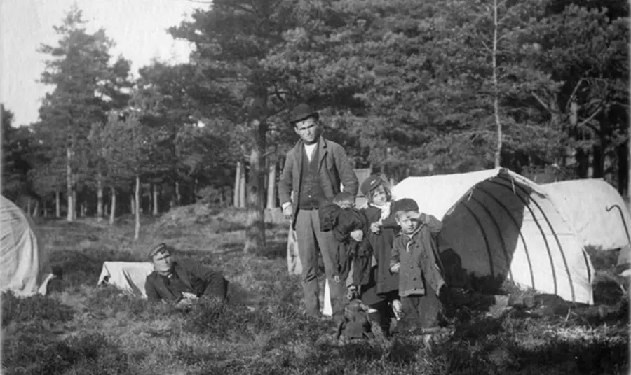 A historic black and white photograph of traveller children and an adult standing in front of tents