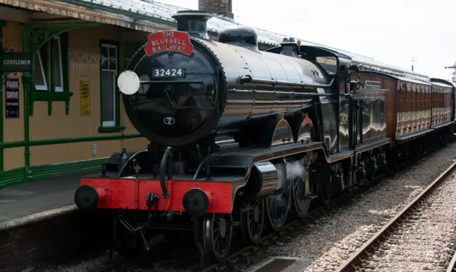 A black steam engine pulling dark green carriages pulls into Horsted Keynes station.