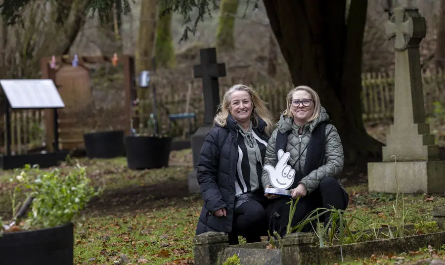 Two people sit with the National Lottery award in Hartwood Paupers Cemetery Hartwood Paupers Cemetery
