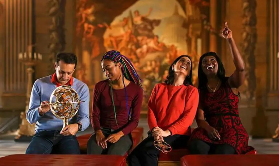 Four people sitting on a bench in a gallery; two looking up towards the ceiling and two looking at a globe one of them is holding