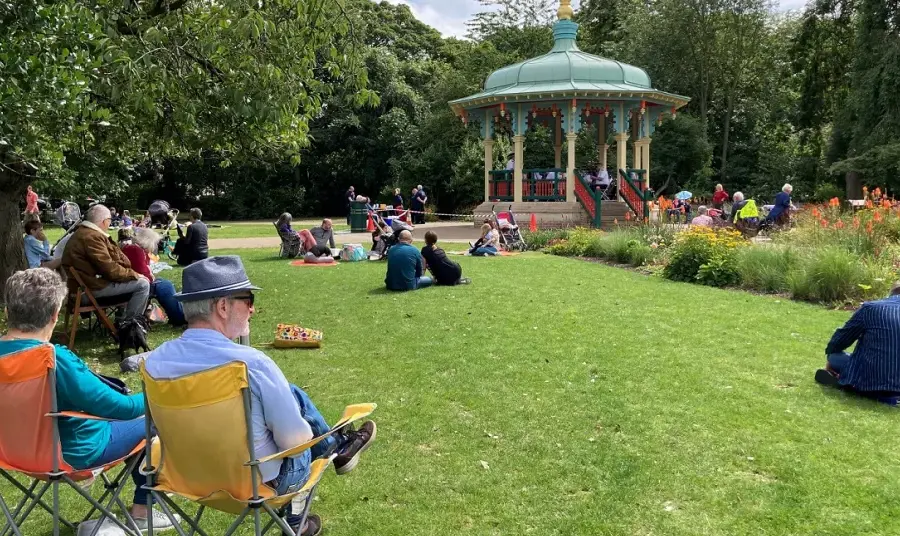 People sitting on camping chairs and on the grass in Pearson park in front of a historic bandstand